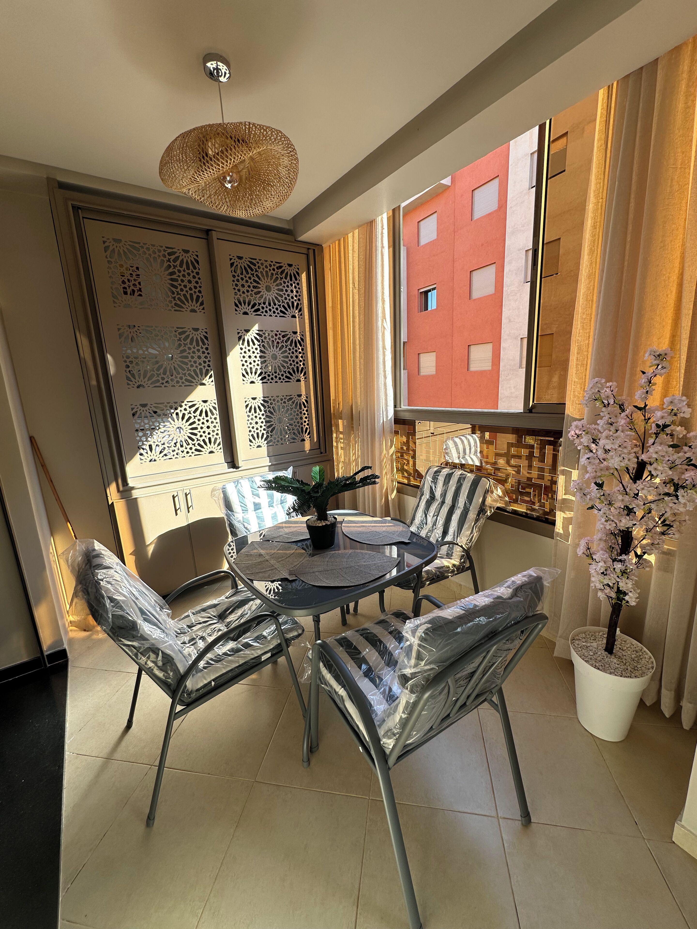 A dining area with a round table and four chairs, a decorative ceiling light, a potted plant, and a view of a building with large windows.