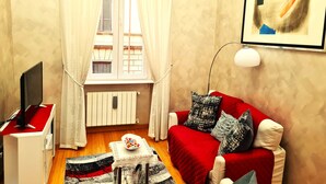Un espace de vie compact avec un canapé rouge, une petite table sur laquelle se trouve un saladier de fruits, un téléviseur posé sur un meuble blanc et une fenêtre avec des rideaux.