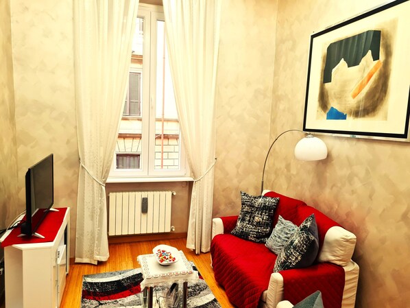 A compact living space with a red sofa, a small table with a bowl of fruit, a television on a white stand, and a window with curtains.