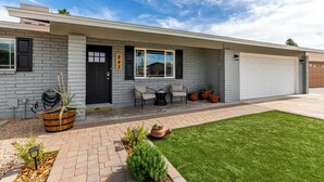 A modern single-story house with a grey brick exterior, a black front door, and a garage. There are outdoor seating with chairs and a table, and potted plants are placed around the entrance.