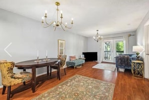A spacious living room with a round wooden table, two chairs, a sofa, a blue armchair, a patterned rug, and two chandeliers.