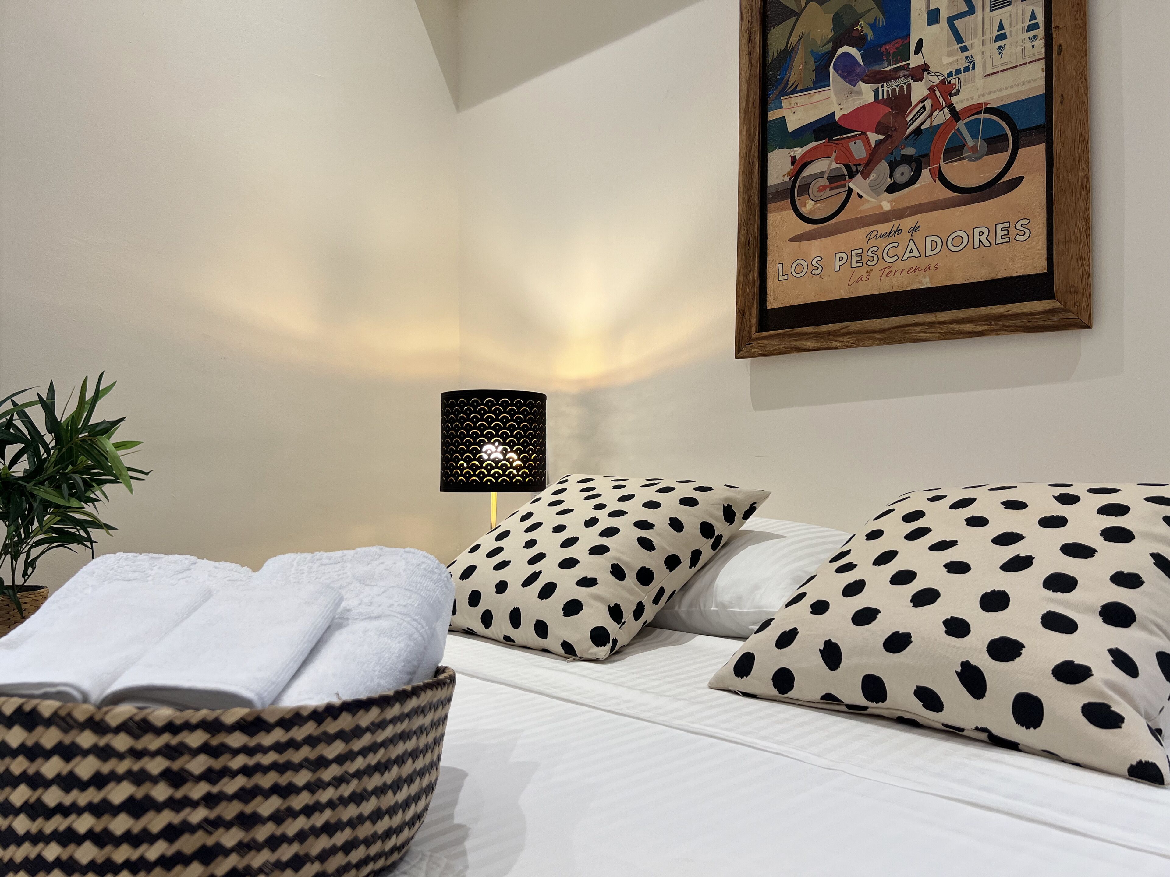 Una cama bien hecha con almohadones de lunares, una cesta tejida con toallas, un cuadro enmarcado en la pared y una lámpara de pie.