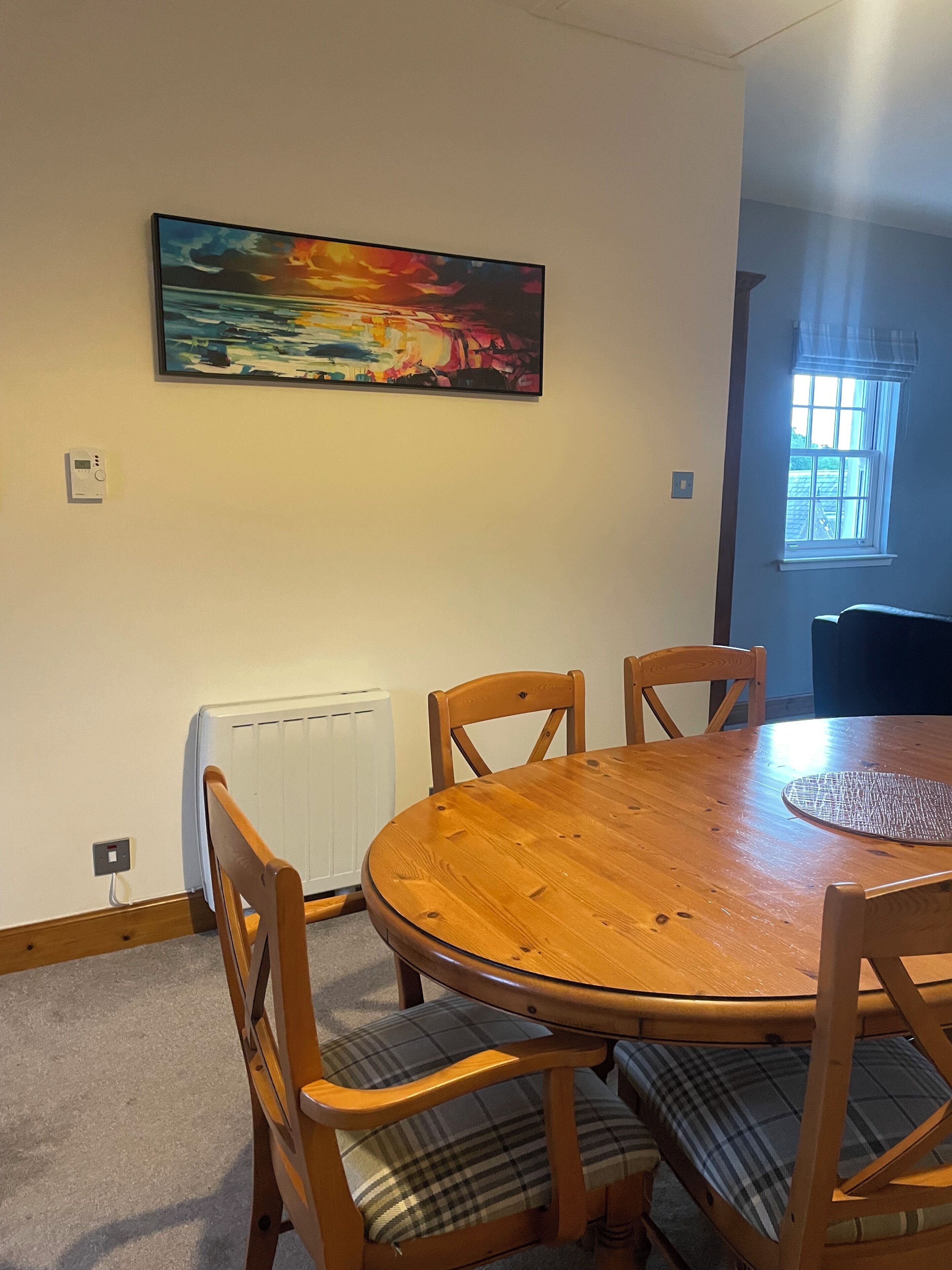 A dining area with a round wooden table, four wooden chairs, a plaid cushion, a painting of a sunset, a window, and a radiator.