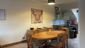 Un coin repas avec une table et des chaises en bois, un tableau représentant une vache accroché au mur, et une cuisine en arrière-plan.