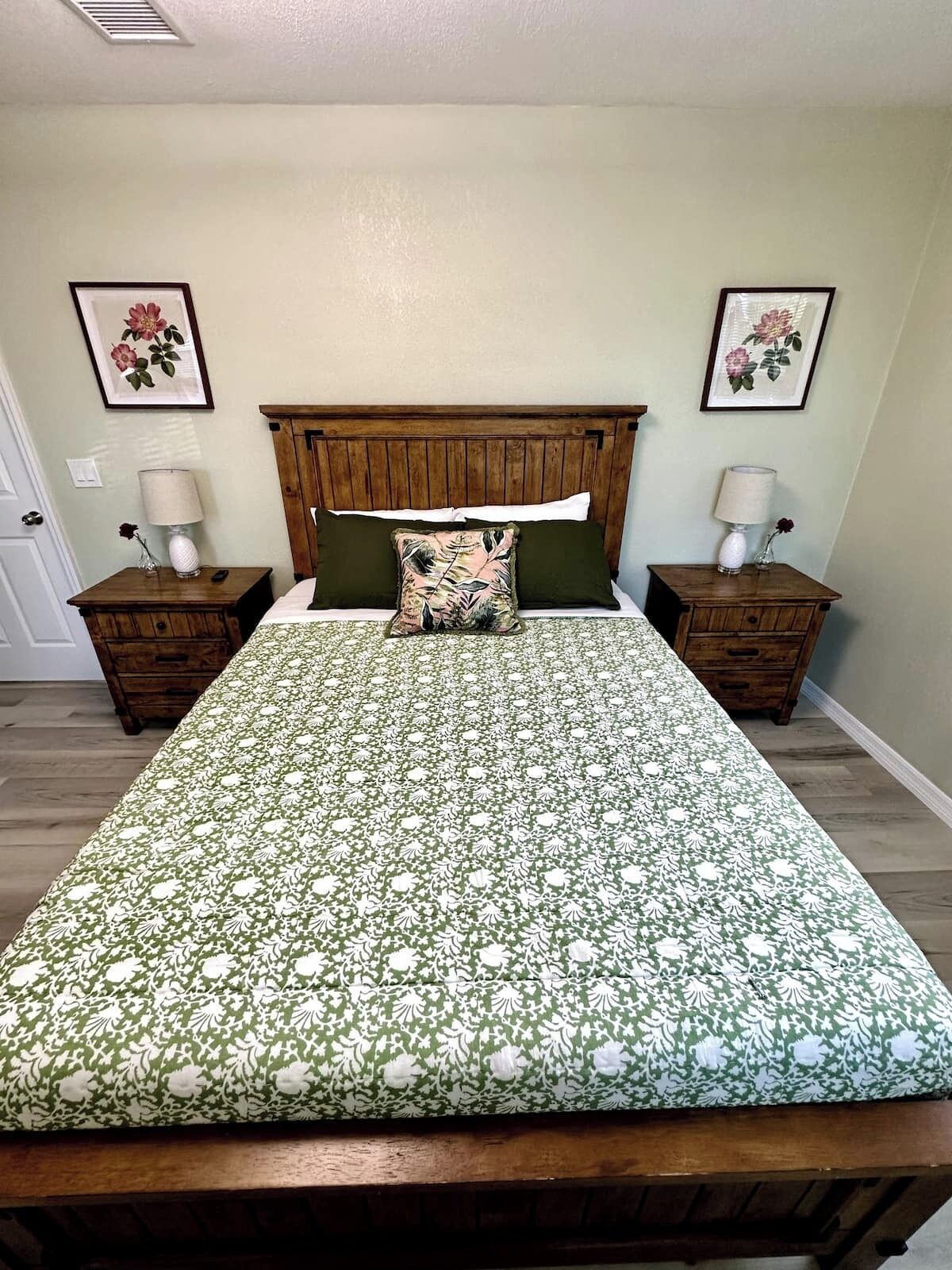 A bedroom with a wooden headboard, a floral bedspread, two nightstands with lamps, and framed floral artwork on the walls.