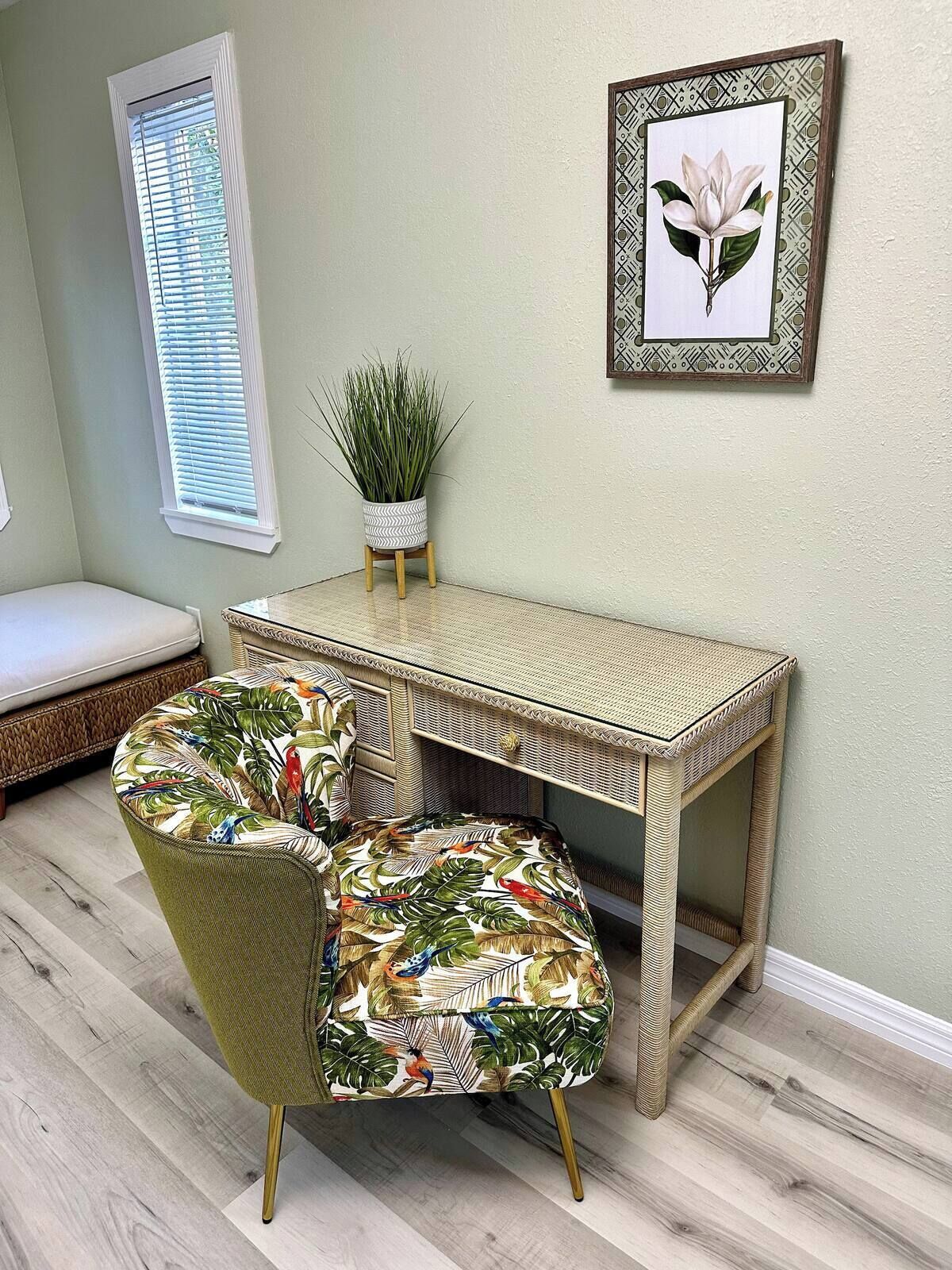 A room with a wicker desk, two chairs with tropical prints, a small potted plant, a framed picture on the wall, and a window with blinds.