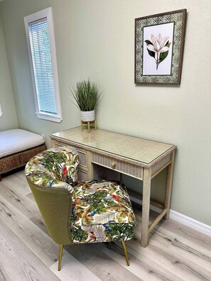 A room with a wicker desk, two chairs with tropical prints, a small potted plant, a framed picture on the wall, and a window with blinds. - Historic District Retreat - Newly Renovated 2BR (Palmetto)