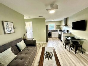 A living room with a sofa, coffee table, and a TV mounted on the wall. There is a kitchen area with a stove and cabinets in the background. - Historic District Retreat - Newly Renovated 2BR (Palmetto)