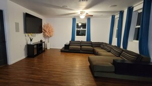 Een woonkamer met een hoekbank, een flatscreen televisie, een plafondventilator en een raam met blauwe gordijnen.