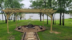 Een houten pergola met een vuurkorf in het midden, omgeven door een grindpad en een trap naar een steiger over het water.