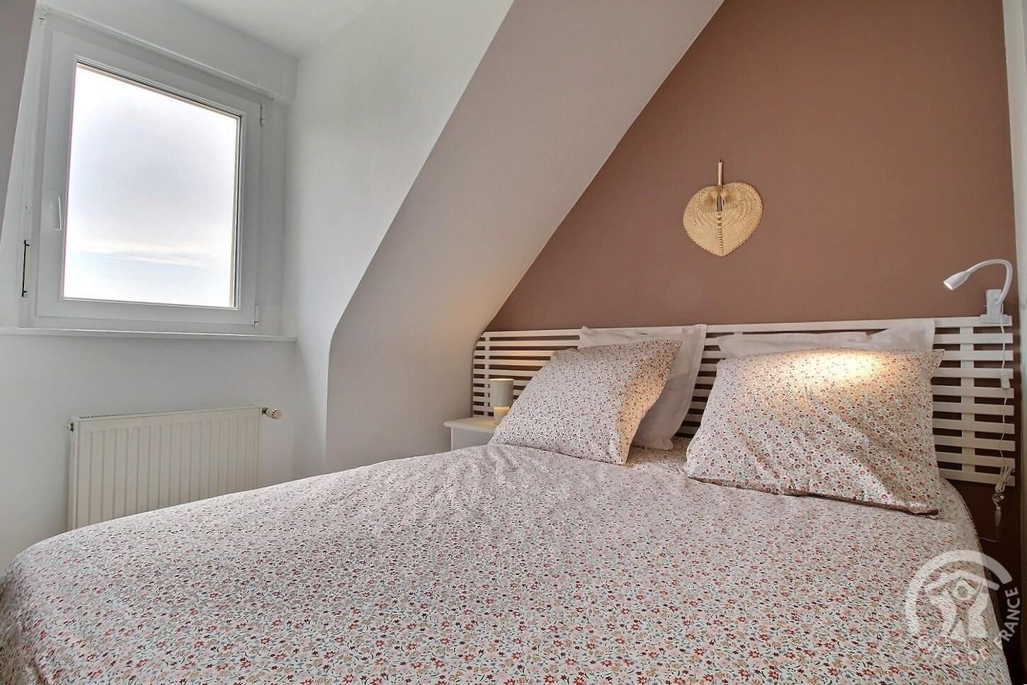 A bedroom with a sloped ceiling, a bed with floral bedding, a bedside lamp, and a window with blinds.