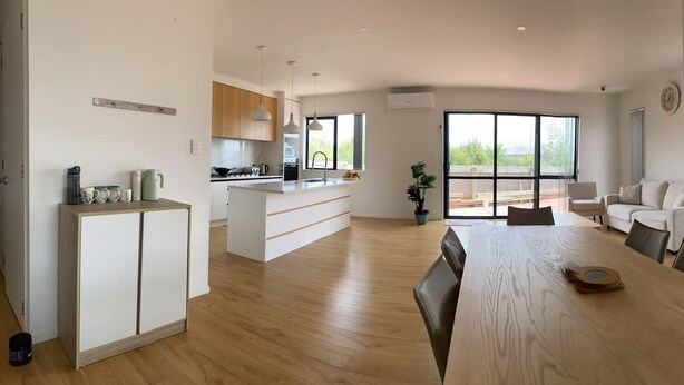 A modern kitchen with white cabinetry, a central island, and pendant lighting. There is a dining area with a wooden table and chairs, and a view of the backyard through large windows.