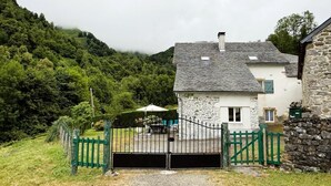 Une maison en pierre à l’extérieur blanc et au toit gris, entourée d’une clôture verte et bordée d’un chemin de gravier menant à une entrée fermée par un portail.