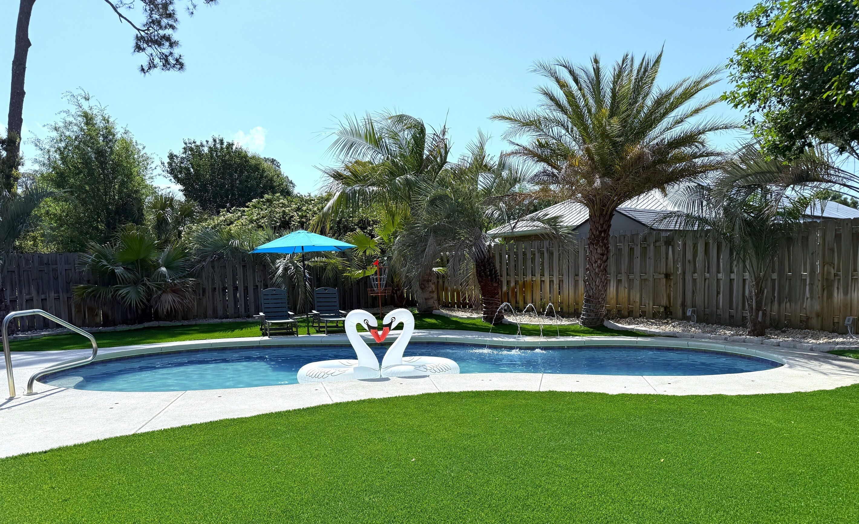 Un patio trasero con piscina, césped artificial, valla de madera y palmeras.