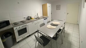 Een kleine keuken met witte kasten, een magnetron, fornuis en een spoelbak. Er is een eettafel met stoelen en een prullenbak.
