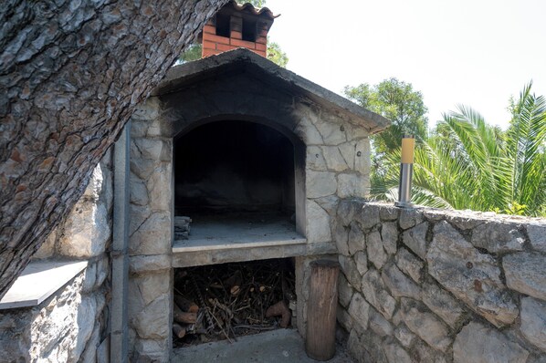 A stone-built outdoor oven with a brick chimney, surrounded by a stone wall and a tree. - Beachfront apartments Nincevic (Trogir)