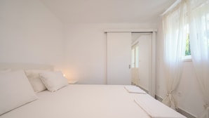 A white room with a bed, a nightstand, and a window with sheer curtains. - Beachfront apartments Nincevic (Trogir)