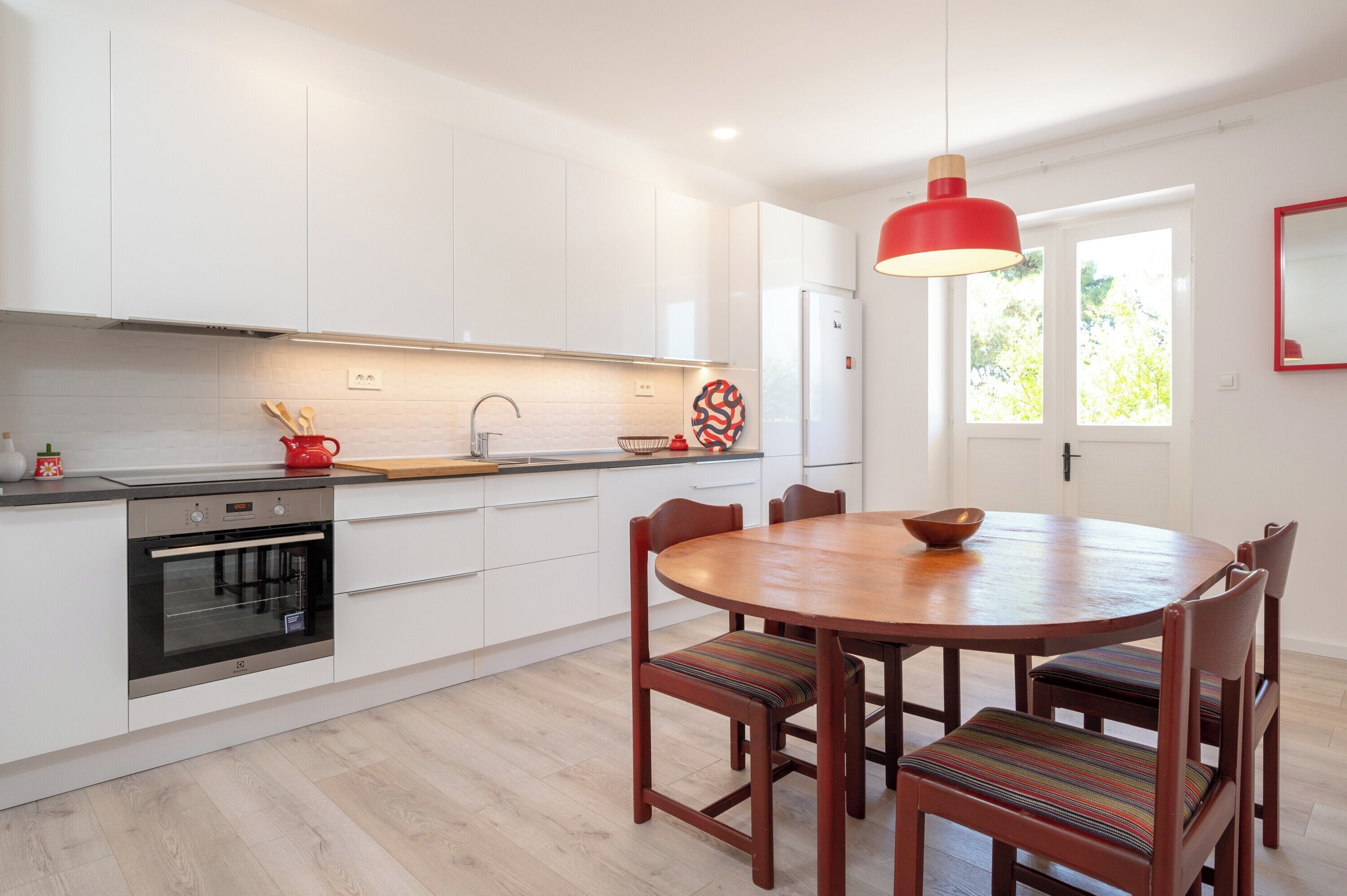 A modern kitchen with white cabinets, a built-in oven, a dining table with chairs, and a red pendant light.