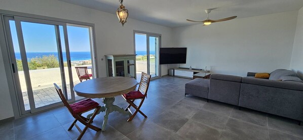 A spacious living area with a large sliding glass door leading to a balcony, a round wooden table with red cushions, a grey sofa, and a ceiling fan.