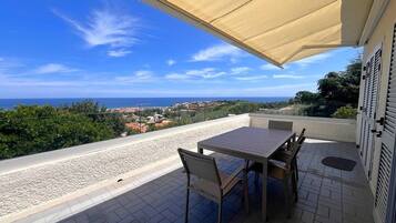 Eine Terrasse mit Tisch und Stühlen, mit Blick auf eine Küstenlandschaft mit Gebäuden und viel Grün.