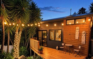 A wooden deck with string lights, a wooden exterior wall, and a glass door leading to an interior space. - Modern Kiama Tiny House - A Unique Coastal Escape (Kiama)