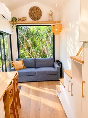 A modern living space with a sofa, dining table, and chairs, a large window with a view of greenery, and a hanging light fixture.