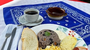 A plate of food including rice, eggs, sausages, and bread, with a cup of coffee and a bowl of soup on a table with a blue tablecloth. - Hotel Cortez Azul (Alajuela)