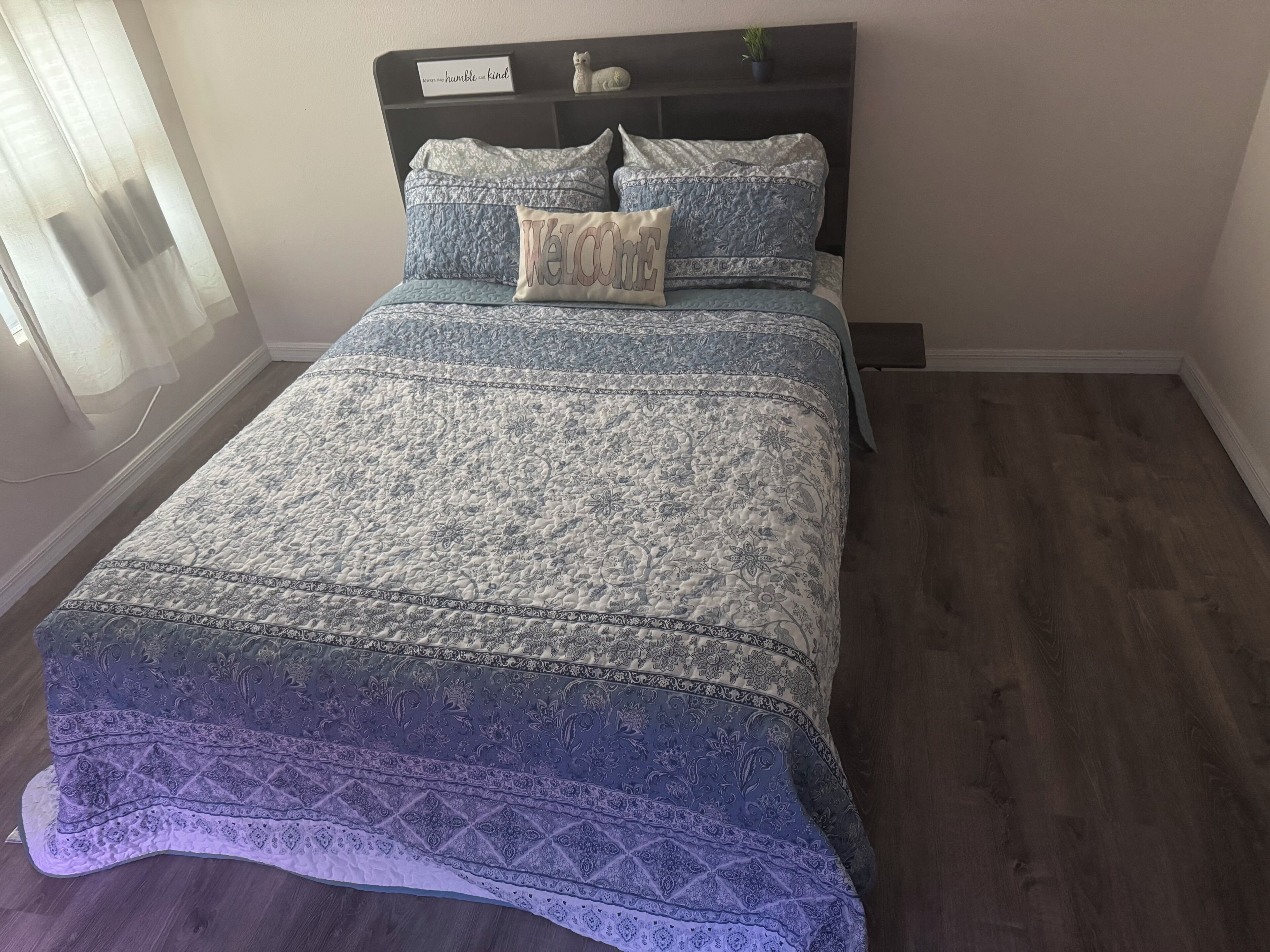 Un lit bien fait, avec une couvre-lit à motifs et des oreillers, une tête de lit sombre et une petite plante posée sur l’étagère au-dessus.