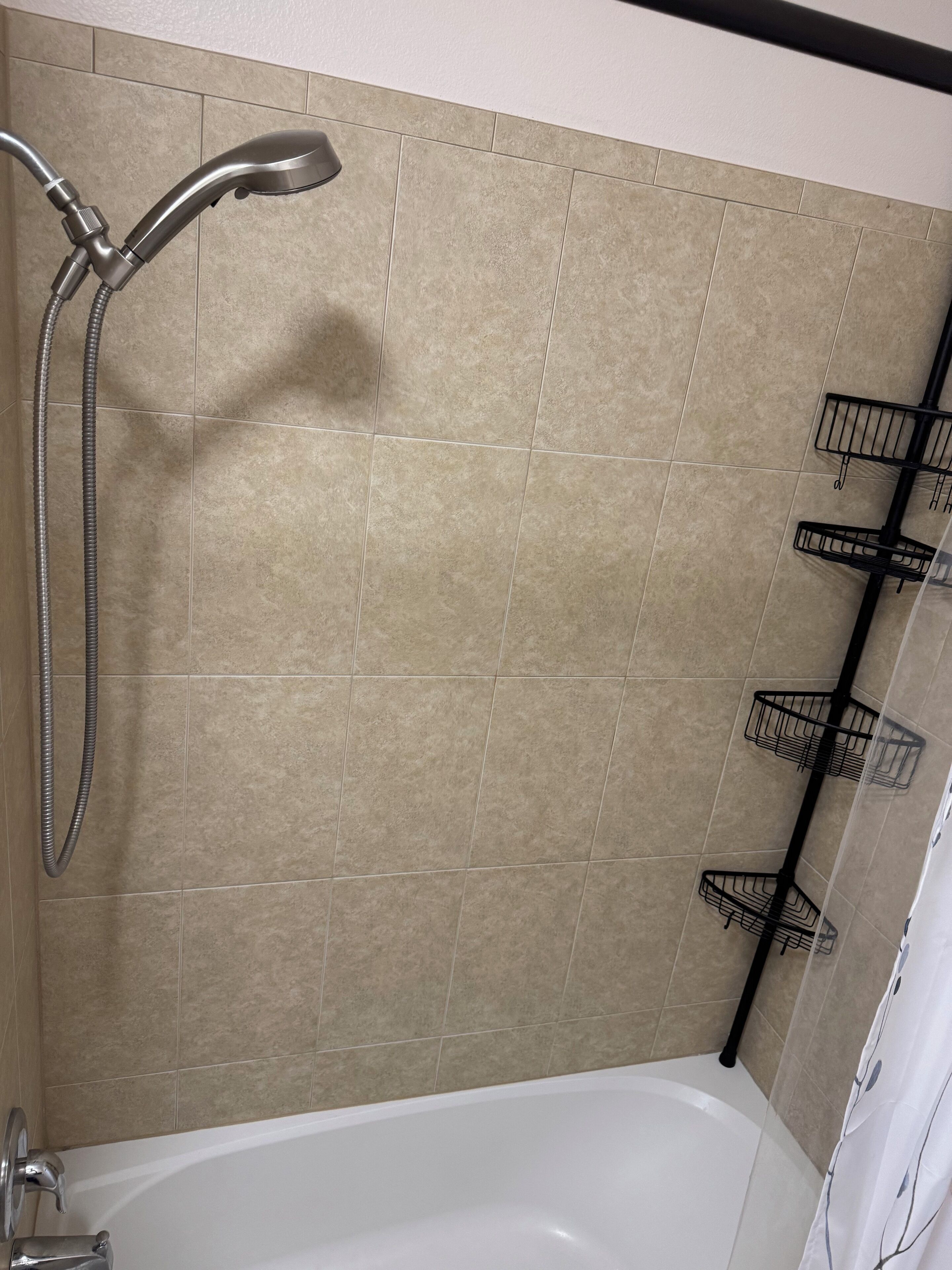 A bathroom with beige tiles, a white bathtub, a shower with a handheld showerhead, and black towel racks.