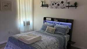Un dormitorio con una cama, ventilador de techo, ventana con cortinas y estantes empotrados con adornos.