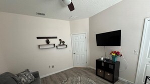Una sala de estar con un sofá gris, un tapete redondo estampado, un televisor de pantalla plana sobre un soporte negro y una mesita con un jarrón de flores.