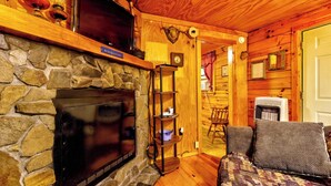 Ein gemütliches Blockhaus mit Kamin aus Stein, Holzwänden, einem darüber hängenden Fernseher und einer komfortablen Sitzecke mit gemustertem Teppich.