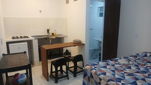 Ein kleines Zimmer mit einem Bett, einem kleinen Schreibtisch mit Stuhl, einer Küchenzeile und einem durch eine offene Tür einsehbaren Badezimmer.