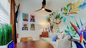 Un salon avec une table ronde en bois, un canapé, un ventilateur de plafond et une peinture abstraite aux couleurs vives accrochée au mur.