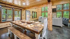 A wooden cabin interior with a dining table set for four, a sofa, and large windows offering a view of the forest.