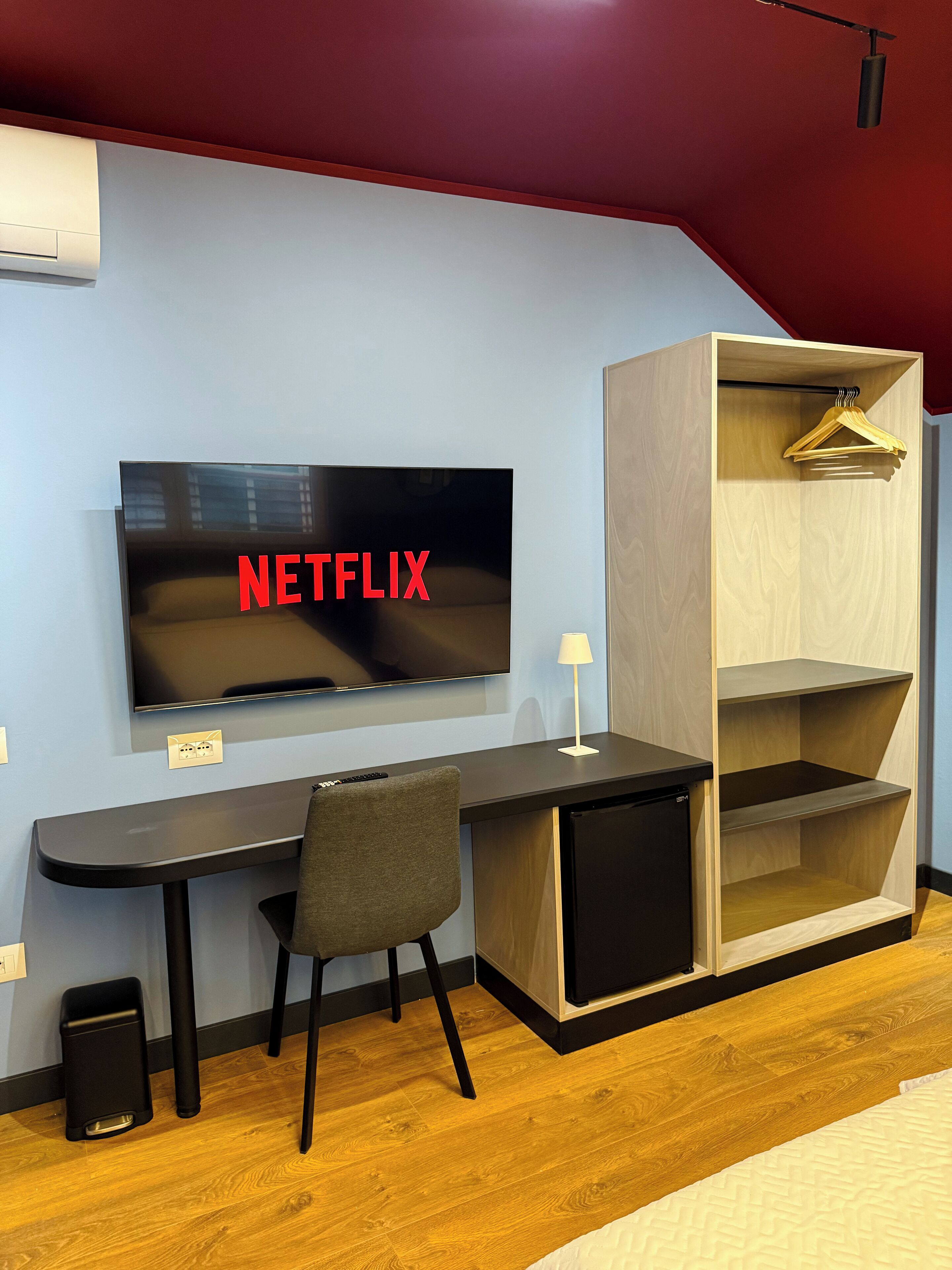 A modern room with a flat-screen TV mounted on the wall, a wooden desk with a chair, a small black speaker, and a wardrobe with shelves.
