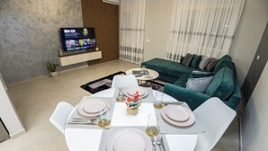 Una sala de estar moderna con comedor listo para cuatro personas. Un televisor de pantalla plana está montado en la pared, y un sofá verde se encuentra al fondo.