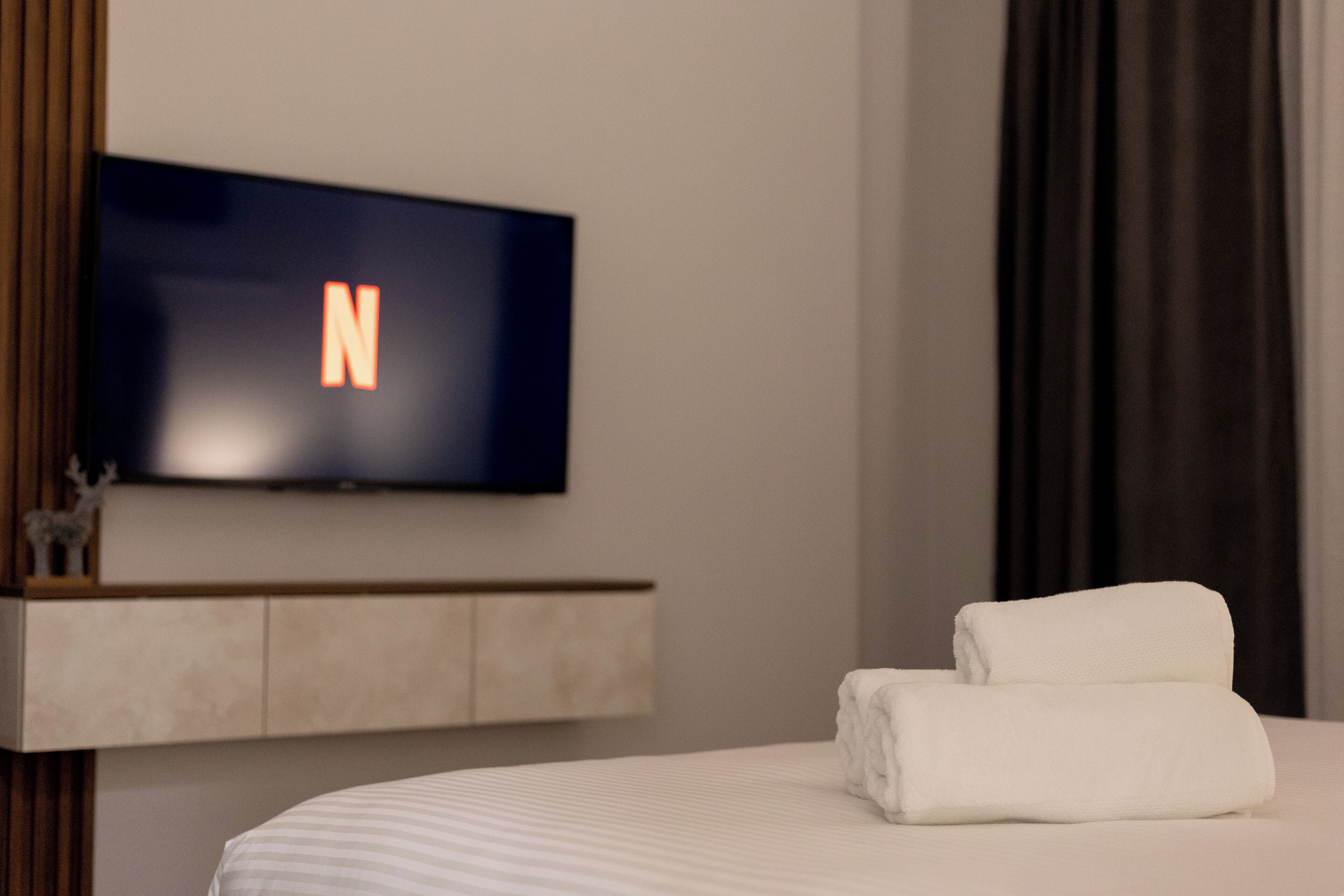 A hotel room with a flat-screen TV mounted on the wall displaying the letter 'N', a neatly made bed with white linens, and a wooden headboard.