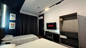 Habitación de hotel con dos camas, un escritorio con un televisor mostrando 'NETFLIX', un armario y obras de arte en las paredes.