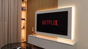Una sala moderna con un televisor de pantalla plana grande montado en una pared blanca, una mesa de centro de vidrio y una mesita auxiliar con un control remoto.