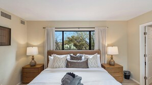 A neatly made bed with a folded towel, two bedside tables with lamps, a window with curtains, and a door.