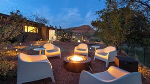 An outdoor seating area with modern chairs and a central fire pit, surrounded by a landscaped garden and a building with large windows.