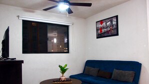 A living room with a blue sofa, a small round table with a welcome sign, a ceiling fan, a window with blinds, and a framed picture on the wall. - Furnished apartment in downtown Guadalajara (Guadalajara)