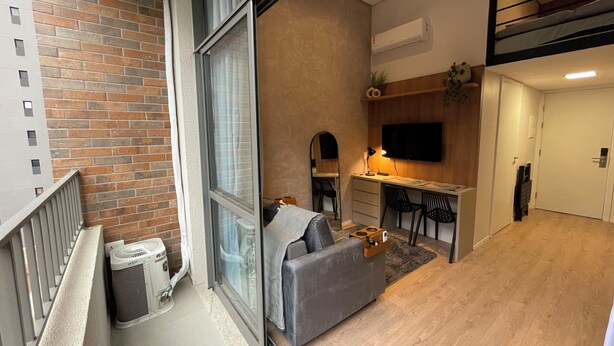 A compact living space with a kitchenette, a grey sofa, a dining area, and a TV mounted on the wall. - Beautiful studio with mezzanine in Brooklyn (Jardim das Acacias São Paulo)