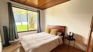 Et soveværelse med træloft, et stort vindue med gardiner, en seng med hvidt sengetøj og to gule puder, samt et lille bord med en lampe og en kop.