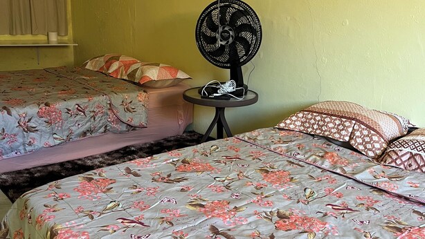 Een kamer met twee bedden, elk voorzien van bloemenbedrukking. Er is een klein tafeltje met een ventilator en een stekkerdoos.