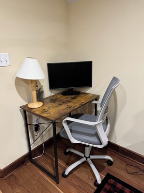 A corner workspace with a wooden desk, a computer monitor, a chair, and a lamp.