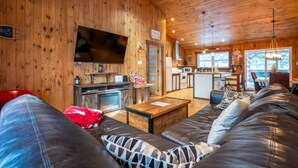A cozy cabin interior with a wooden interior, a large sofa, a television, a fireplace, and a kitchen area. - Chalet Le Shack Sur la rouge (La Conception)