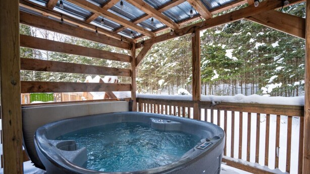 A hot tub in a wooden gazebo with a metal roof, surrounded by snow-covered trees. - Chalet Le Shack Sur la rouge (La Conception)