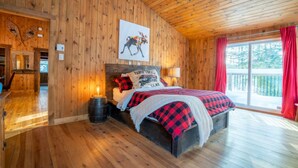 A wooden cabin bedroom with a bed, a nightstand, a wall-mounted painting, and a window with curtains. - Chalet Le Shack Sur la rouge (La Conception)